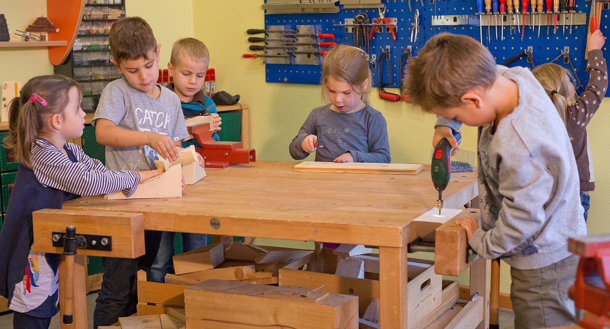 Kinder stehen um einen Holztisch, halten Werkzeug in der Hand und werkeln