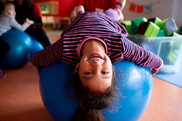 Ein Mädchen liegt auf einem Gymnastikball und lacht kopfüber in die Kamera